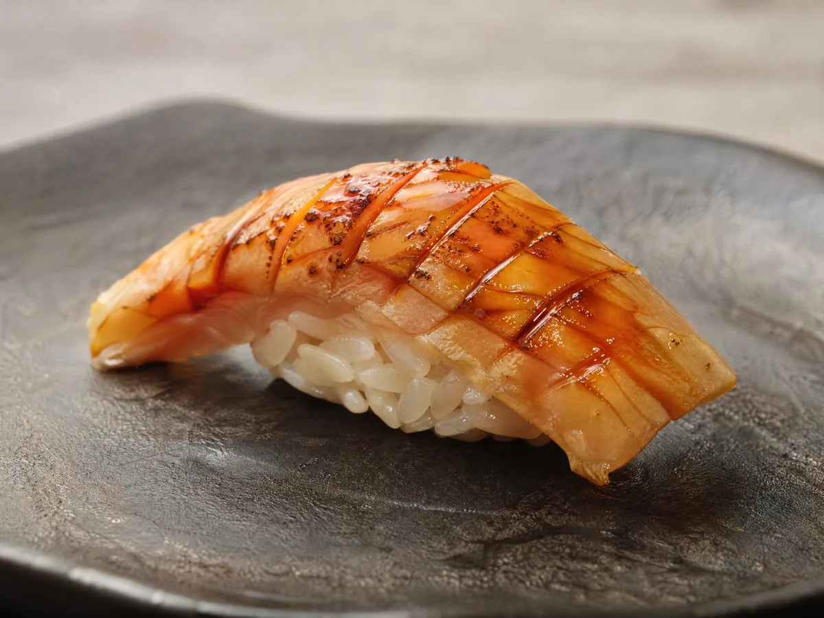 A single piece of scored, glazed nigiri on rice on a dark plate