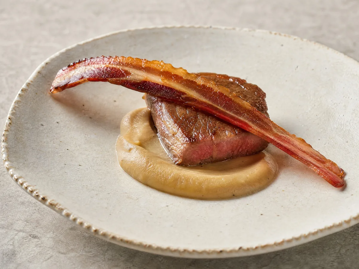 A small seared slice of wagyu on a purée with a strip of bacon, on white china