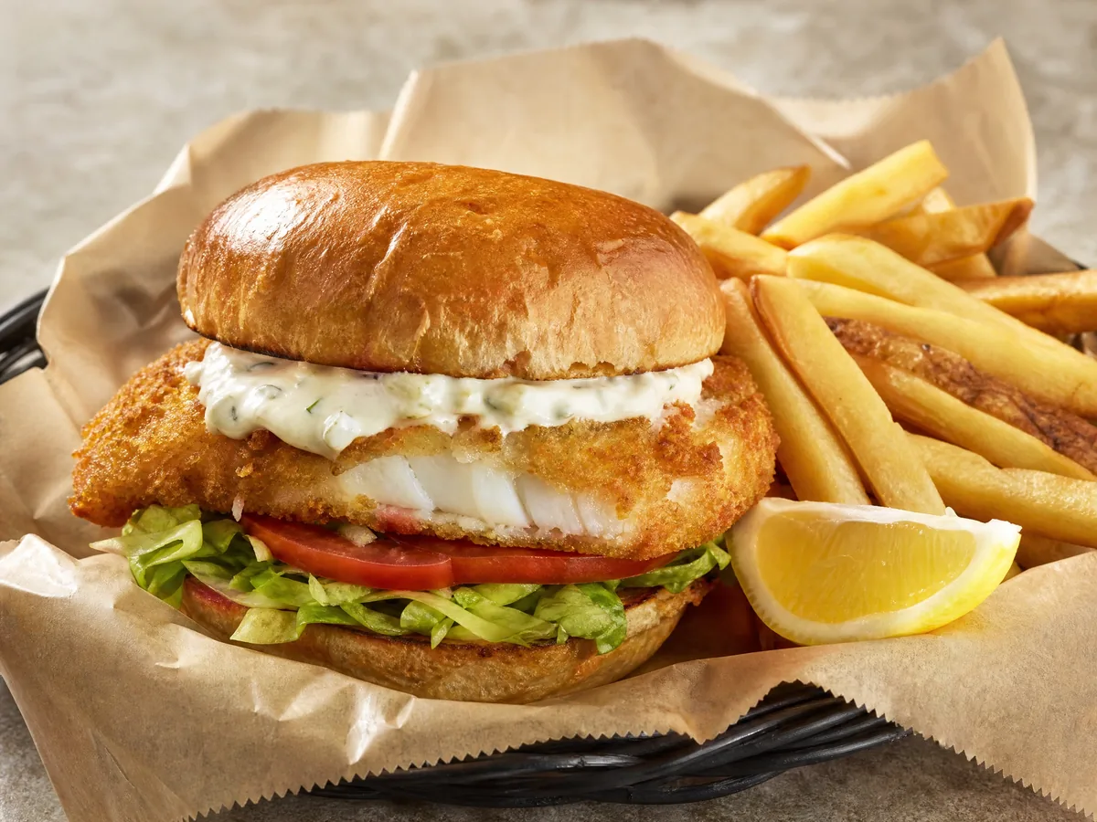 A fried fish sandwich with lettuce, tomato and tartar sauce, with fries and lemon in a basket