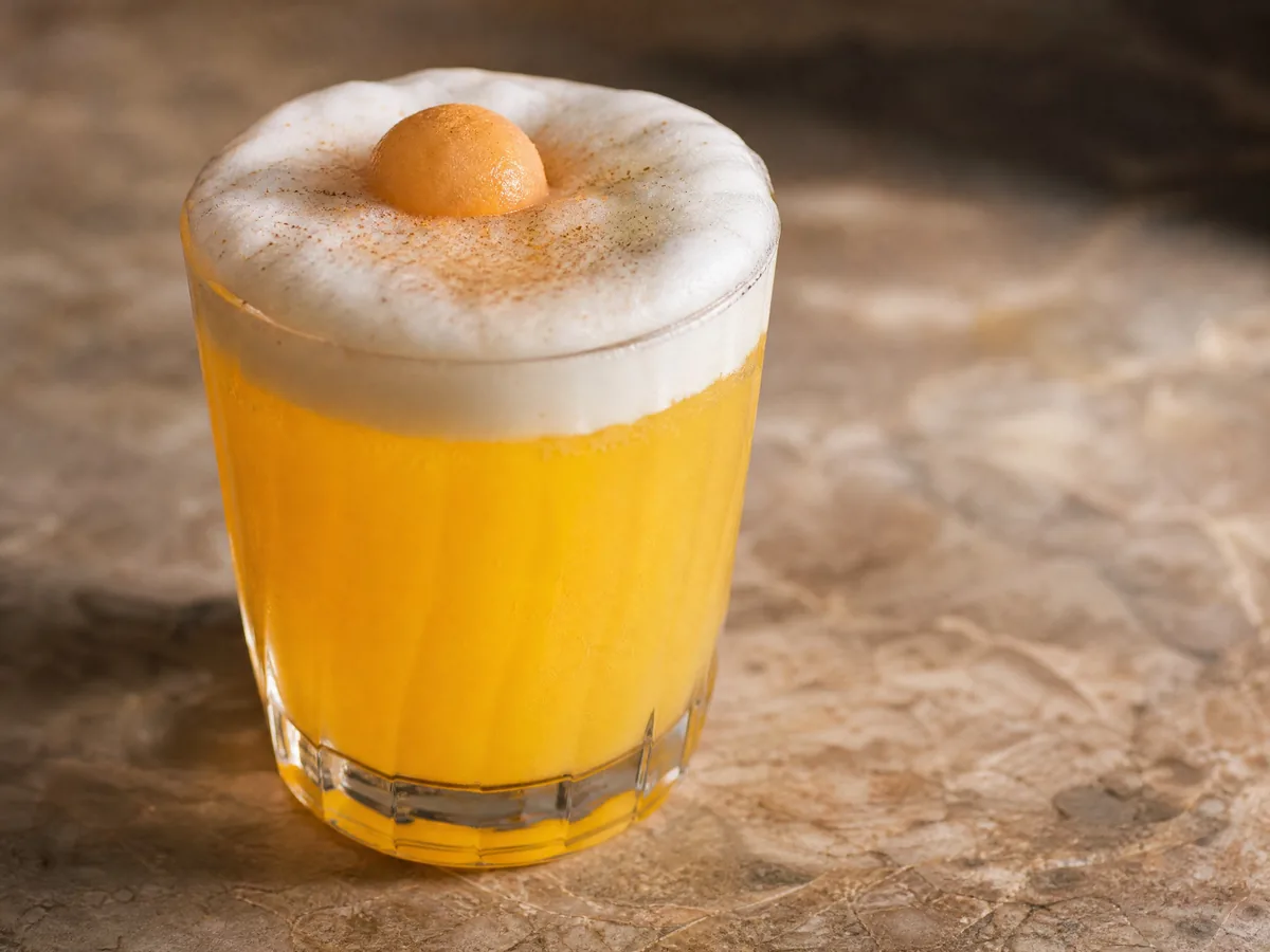 A tall orange cocktail topped with pale foam