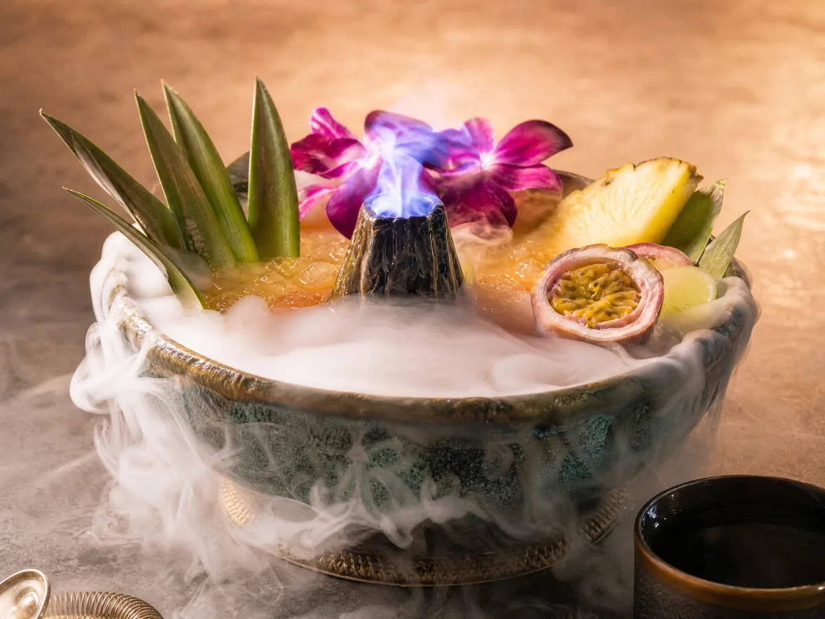 A large tiki bowl drink garnished with orchids and fruit, with cold vapor spilling over the rim