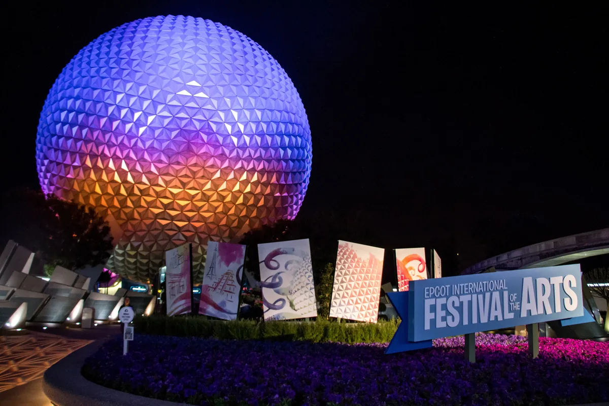 Spaceship Earth lit purple at night above art banners and the Festival of the Arts sign at EPCOT