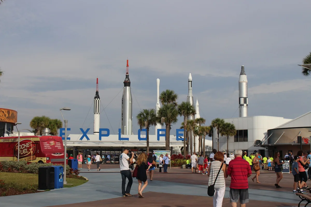 Visitors approach the EXPLORE entrance at Kennedy Space Center with rockets rising behind palms