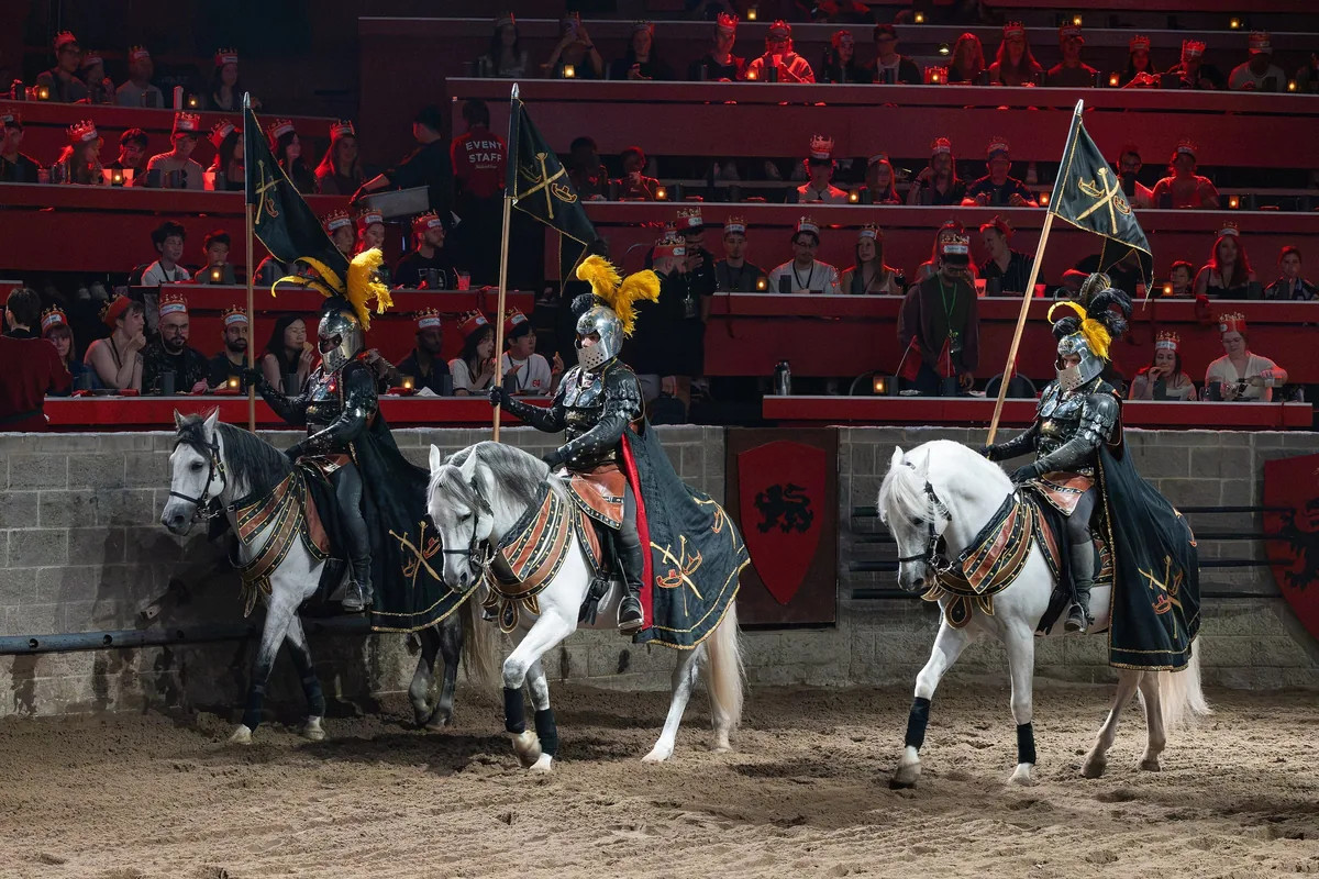 Armored knights on white horses in a Medieval Times sand arena before a crowd of diners