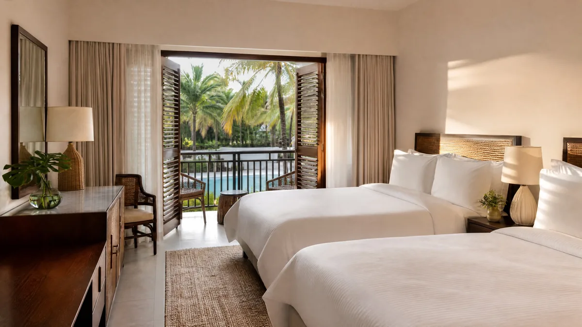 A bright hotel room with two queen beds and an open balcony door onto palm trees and a pool
