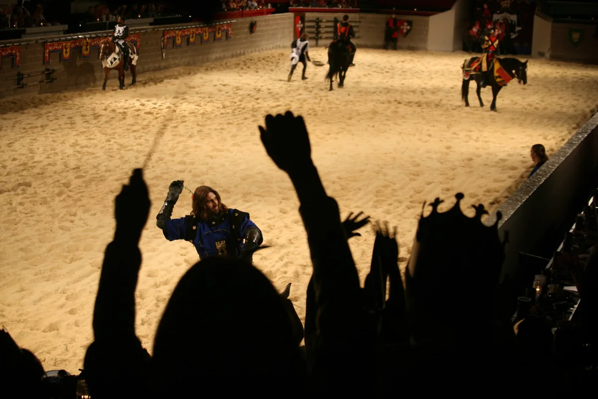 Silhouetted diners cheering as knights on horseback circle a sand jousting arena
